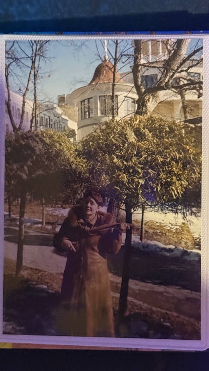 Фотография мамы в длинном пальто на улице, рядом с деревьями и зданием с башенкой